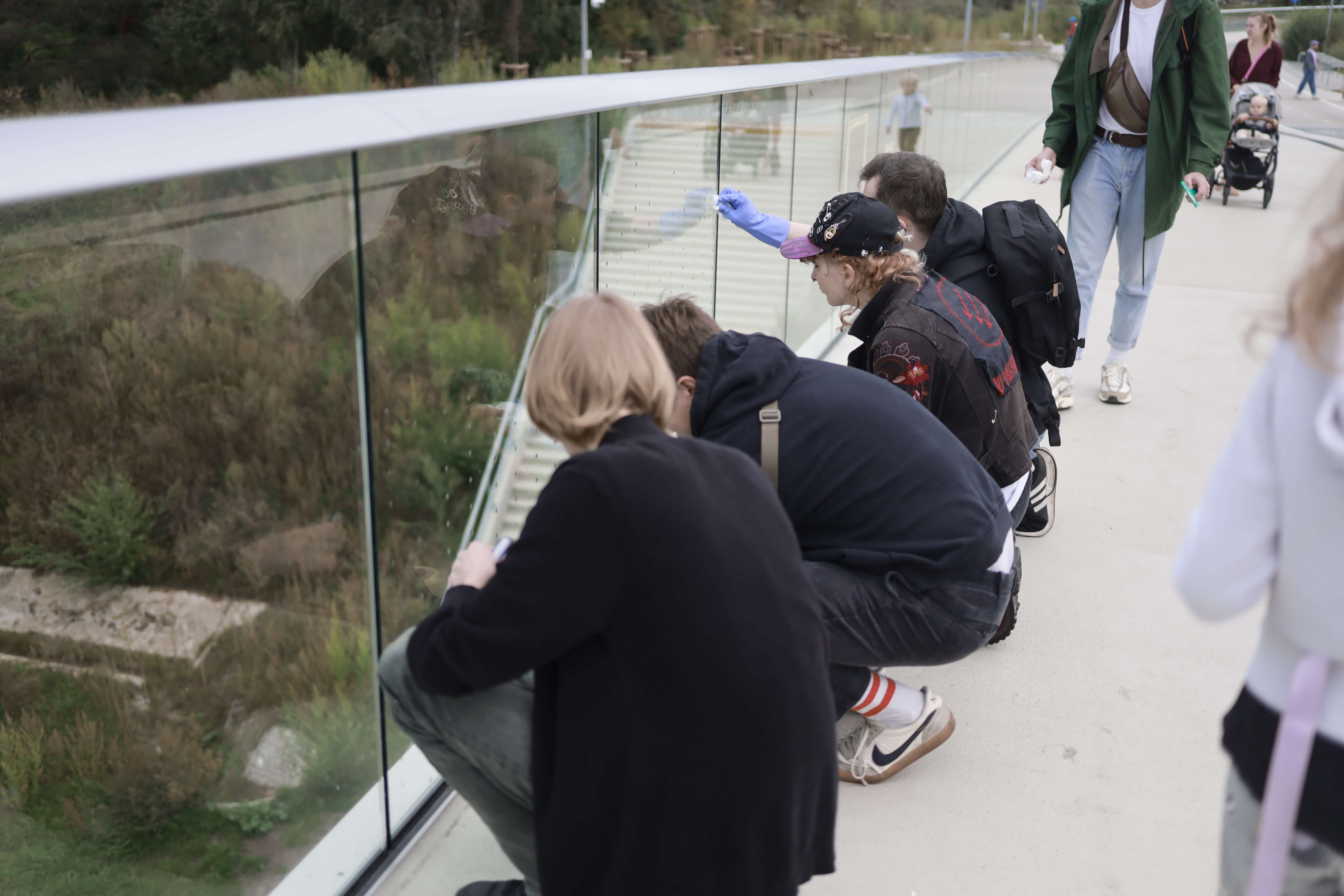 Grupa osób pochyla się przy szklanej balustradzie mostu. Jedna osoba w rękawiczce rysuje lub zaznacza coś na szybie, pozostali obserwują. W tle przechodnie i spacerująca rodzina z wózkiem dziecięcym.